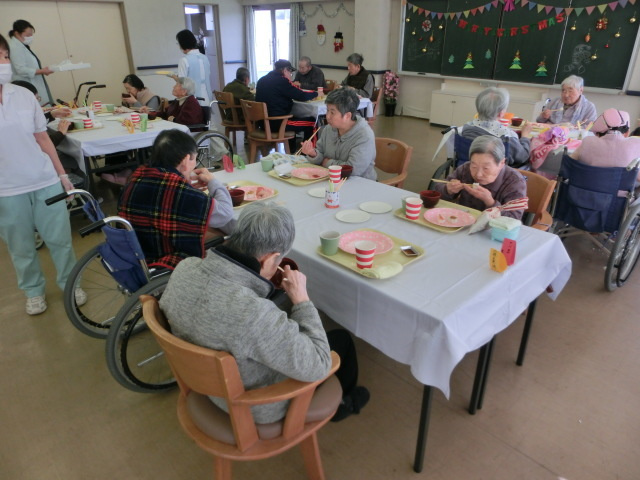 介護施設でのクリスマス時季の食事会。高齢入居者が複数のテーブルで食事と交流を楽しむ様子。